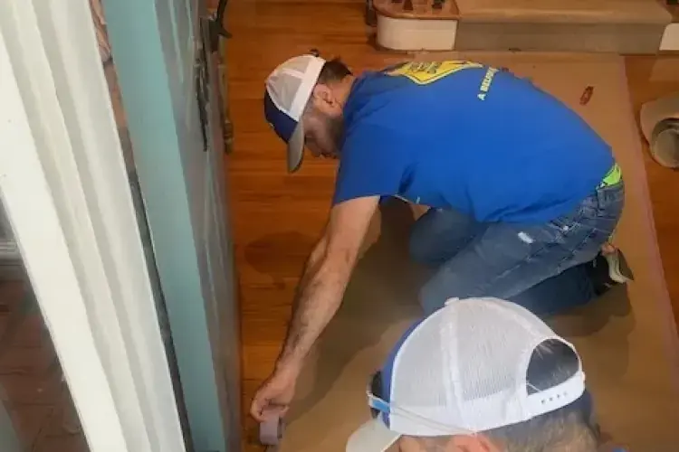 Two workers kneeling on a wood floor checking water damage at the baseboard